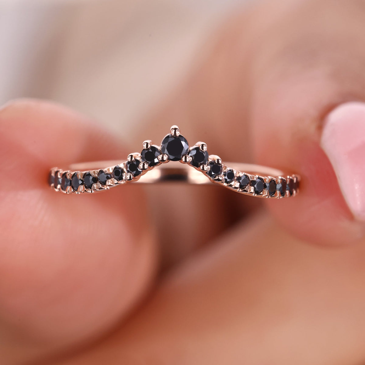 An image of a curved black moissanite wedding band in yellow gold, featuring round black stones in a graceful V-shaped design, held between two fingers.